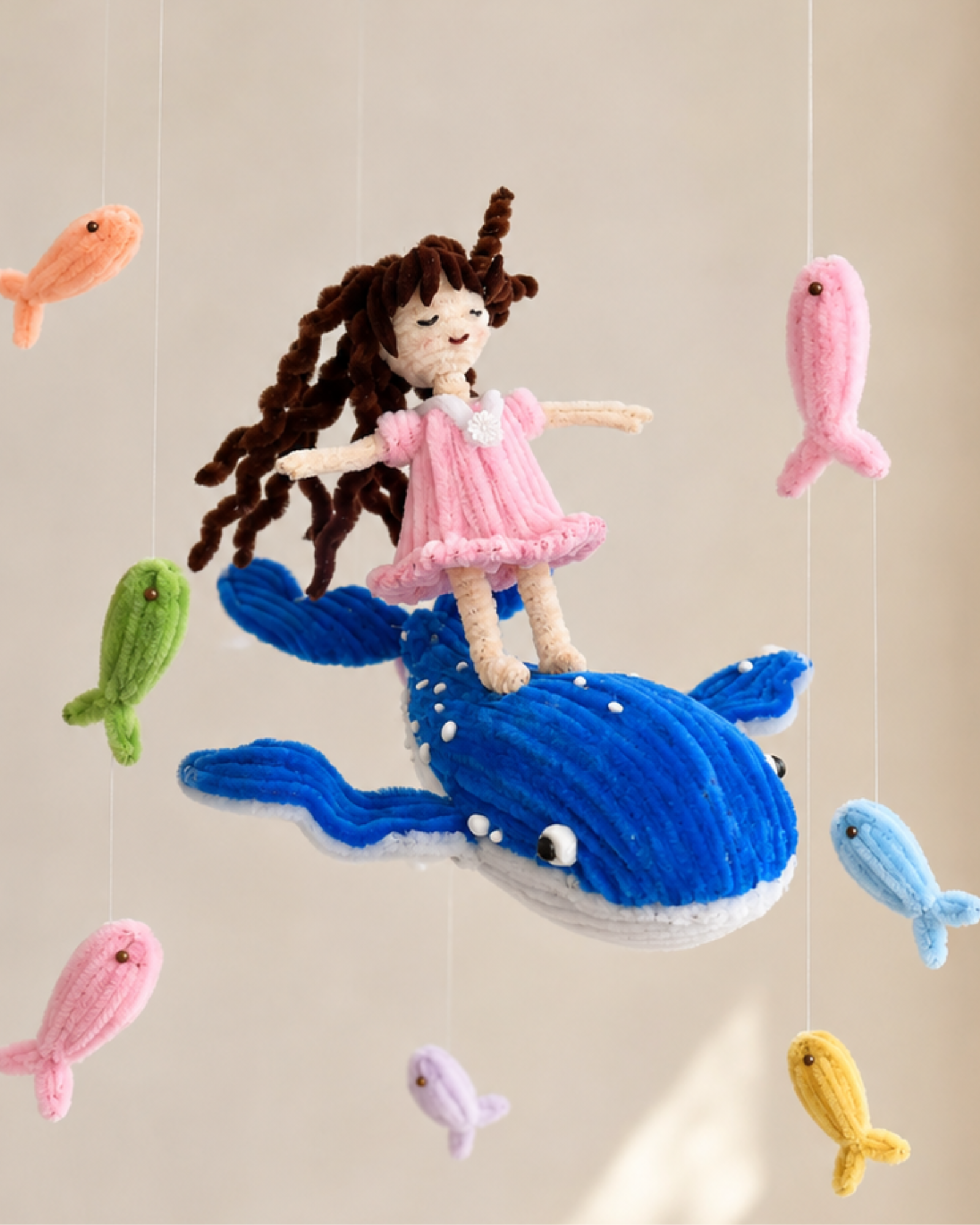 A colorful hanging baby mobile with a blue ring top, small fish charms, and a mermaid figure on a whale, displayed in a bright nursery setting with flowers and a window.