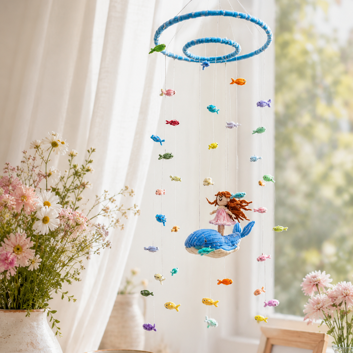 A colorful hanging baby mobile with a blue ring top, small fish charms, and a mermaid figure on a whale, displayed in a bright nursery setting with flowers and a window.